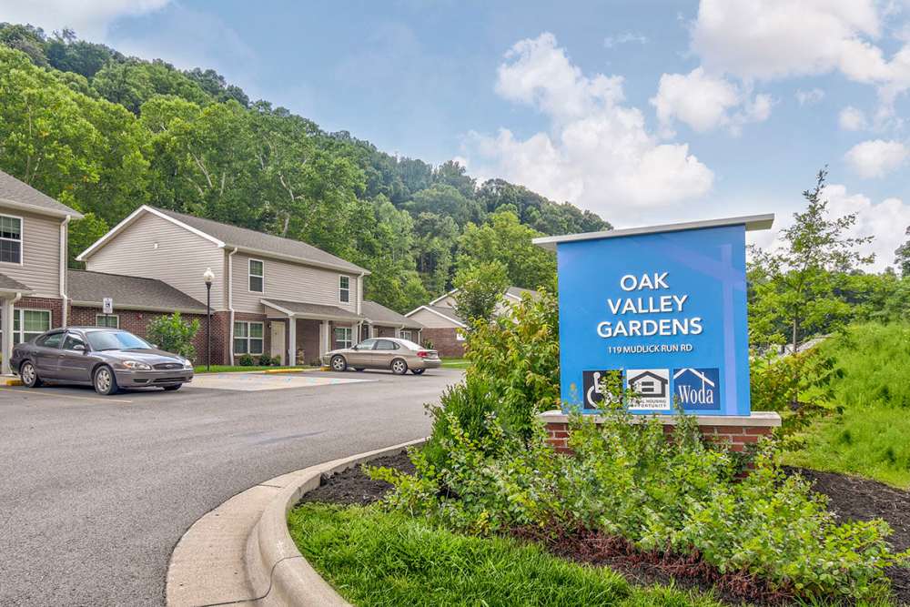 Apartment community entrance with landscaped greenery and signage, two parked cars visible, surrounded by trees at Oak Valley Gardens in Glenville, West Virginia