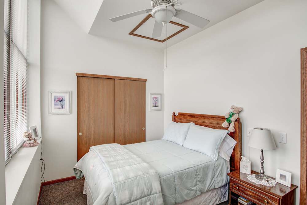 Cozy bedroom with a ceiling fan, wooden bed frame, and light bedding, featuring large windows and closet at Sycamore House Senior Living in Durand, Michigan
