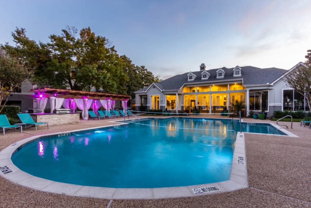 Pool with volleyball net, clubhouse, and colorful lights lounge area at sunset at Marquis at Stonegate in Fort Worth, Texas