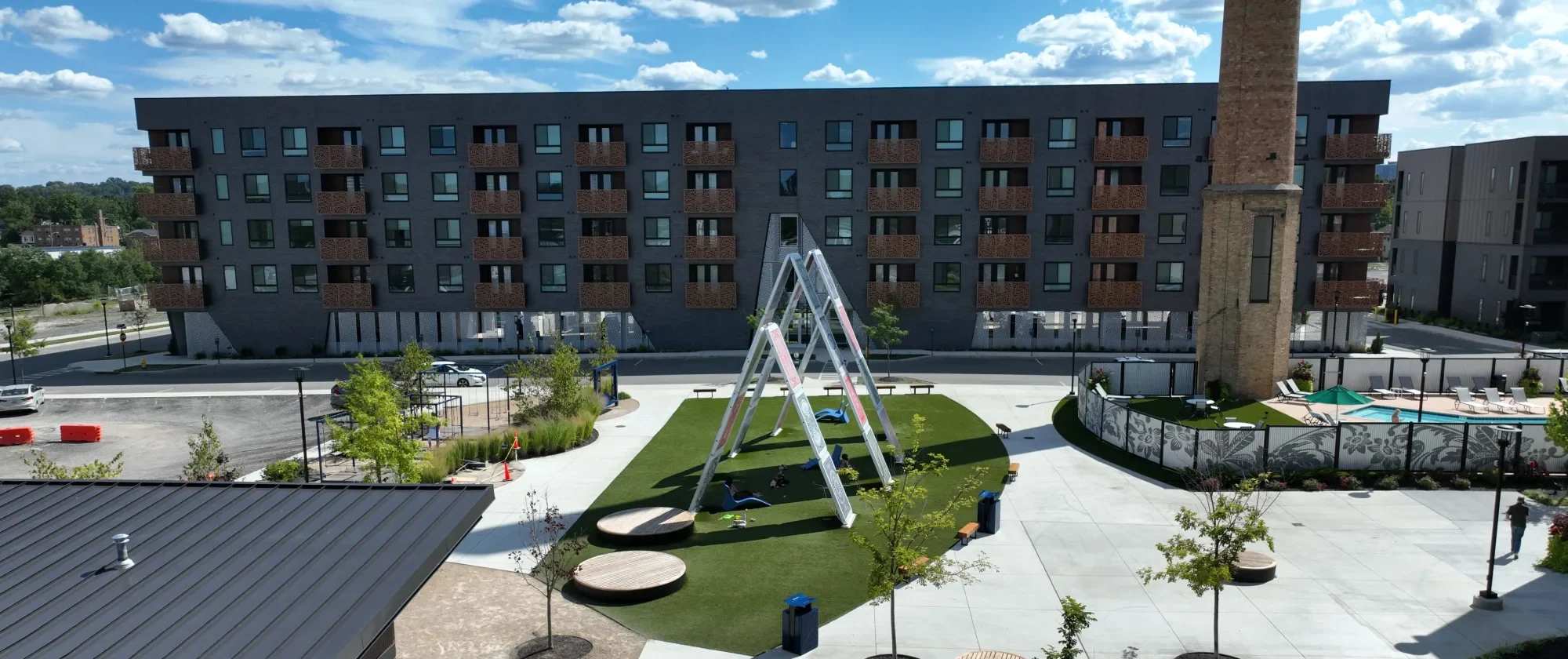 Factory 52 Phase Two apartment building with modern façade, private balconies, central green space, playground structure, and adjacent pool area under a bright blue sky.