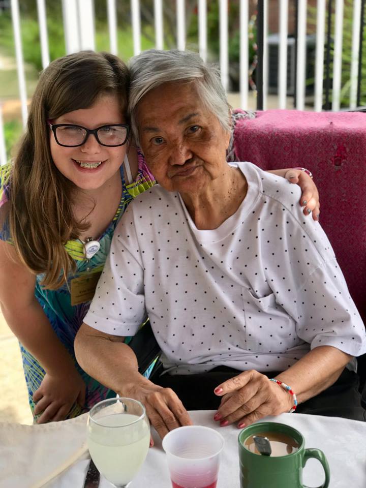 A young woman smiles with her arm around an elderly woman who is enjoying a cup of tea on a sunny afternoon.
