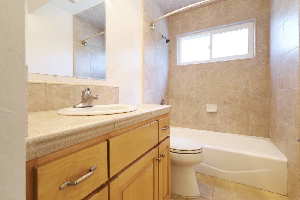 Bathroom with wooden vanity, sink and mirror, toilet, and bathtub with tiled walls and a window above allowing natural light. at Santa Cruz Coast Homes in Santa Cruz, California