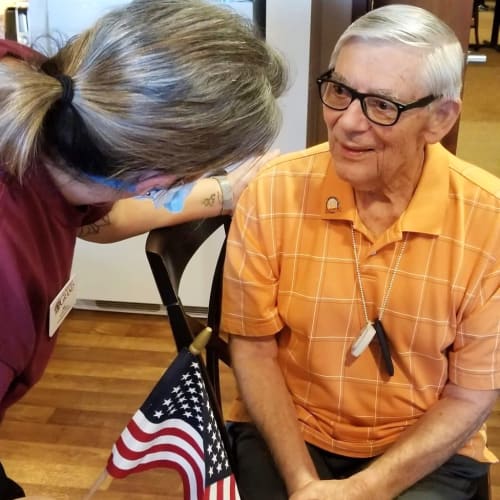 Masked caretaker talking to a seated resident at The Oxford Grand Assisted Living & Memory Care in Wichita, Kansas