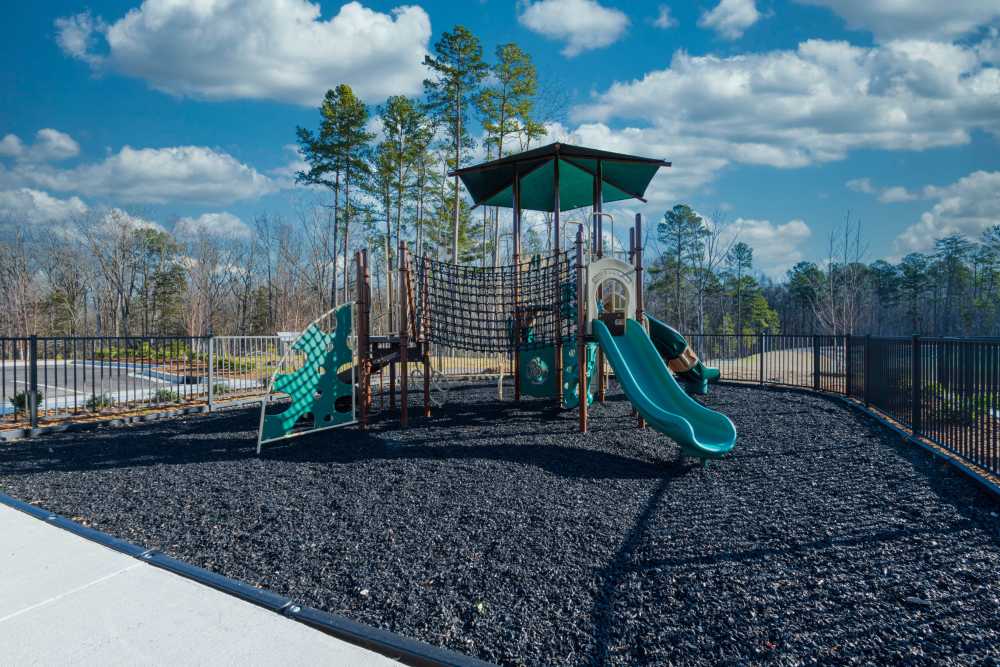 Outdoor playground featuring slides, climbing structures, and safety flooring enclosed by fencing with trees in the background at The Reserve at Patterson Place in Durham, North Carolina.
