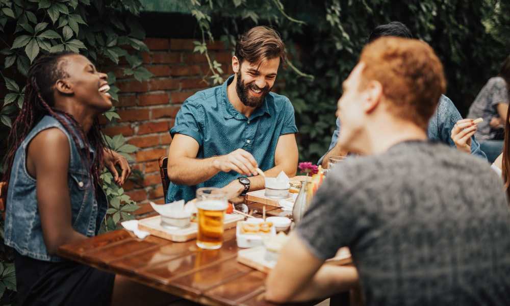 Group of friends laughing and enjoying food at a wooden table surrounded by greenery at Everett at Port Wentworth in Port Wentworth, Georgia