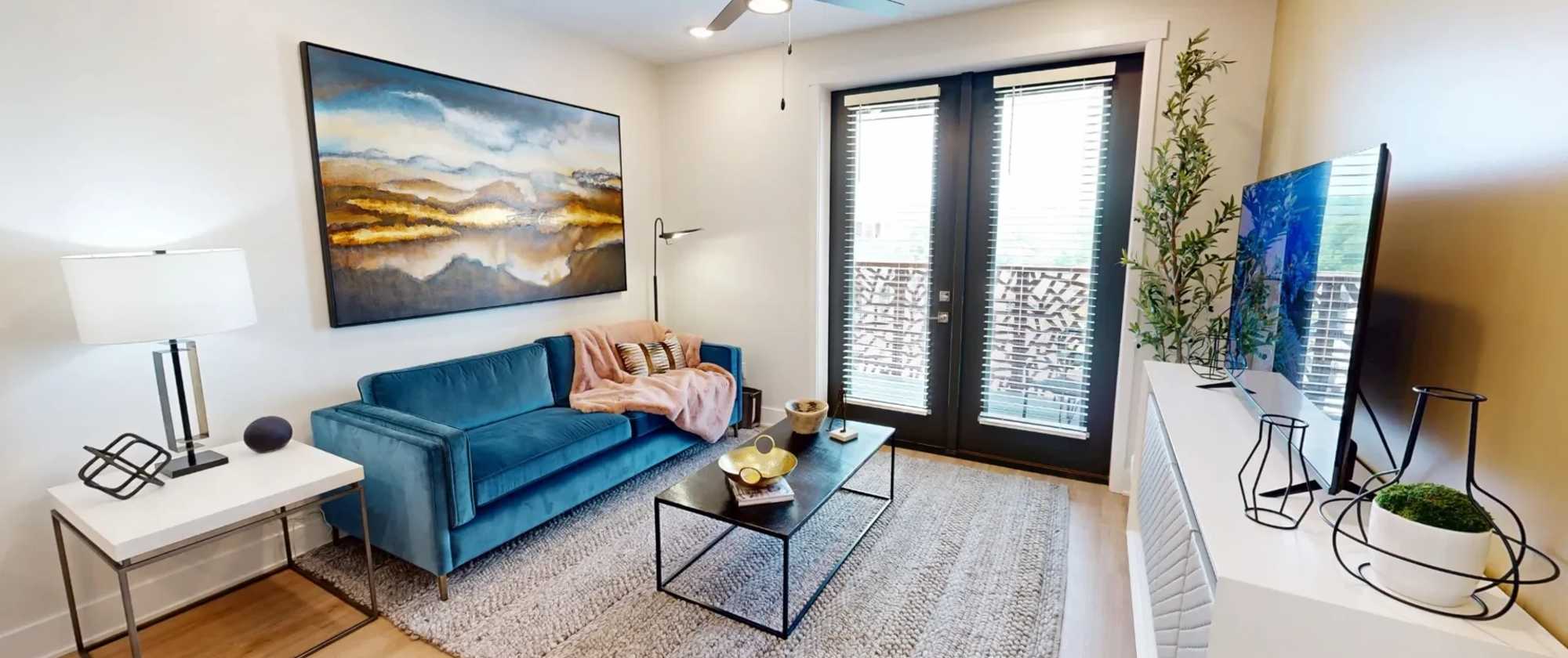 Modern living room inside the Heart II floor plan at Factory 52 Phase Two, featuring a blue velvet sofa, large wall art, natural light from balcony doors, and contemporary décor.