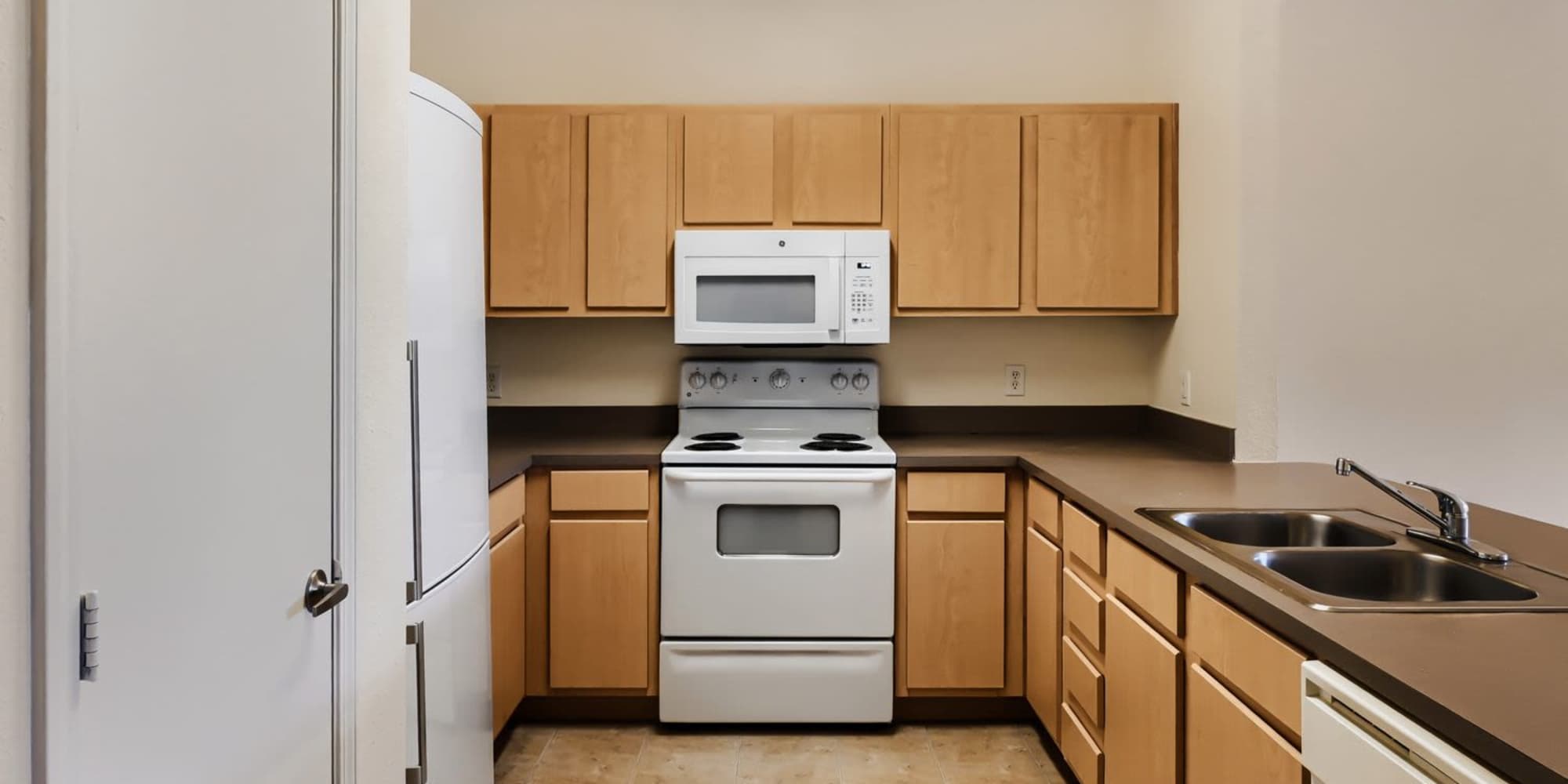 Modern kitchen with beige cabinetry, white appliances, and double sink on a brown countertop at Lindbergh Parc Senior Apartments in Fort Worth, Texas