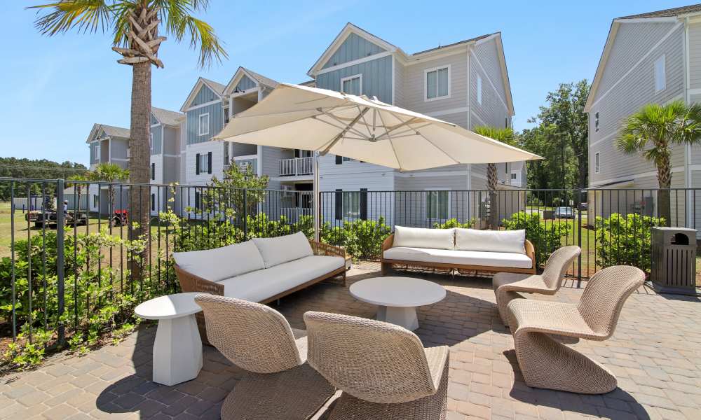 Outdoor lounge area with modern furniture, shaded by an umbrella, surrounded by greenery and palm trees at Everett at Port Wentworth in Port Wentworth, Georgia