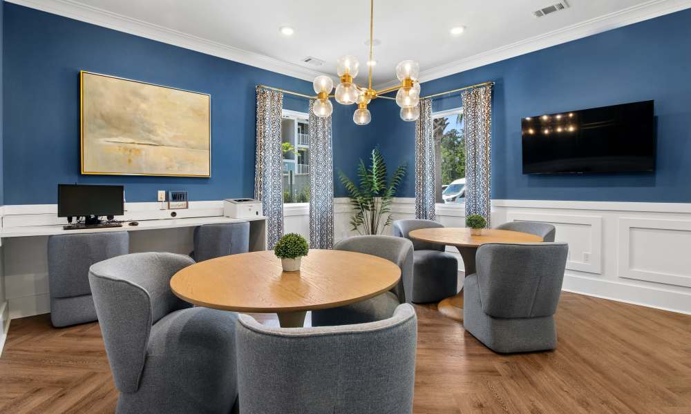 Modern community room with wood-style flooring, round tables, and stylish seating, featuring a large TV and computer station at Everett at Port Wentworth in Port Wentworth, Georgia