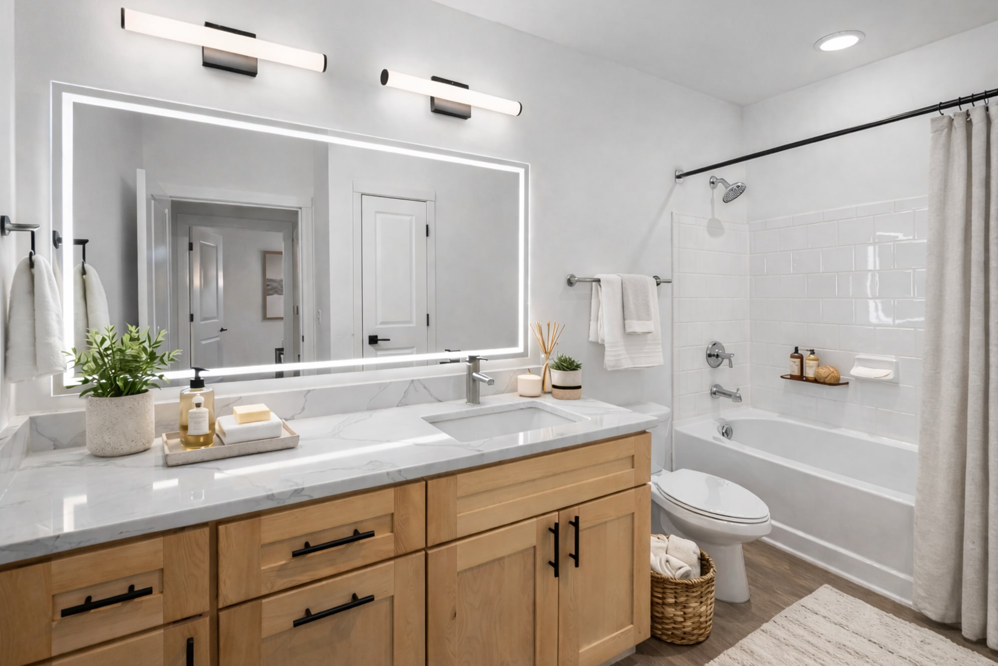 The Aaron 1-bedroom floor plan bathroom at The Carillon in Nashville, TN featuring a light wood vanity with quartz-style countertop, oversized LED mirror, matte black fixtures, and a tub-shower with tile surround.