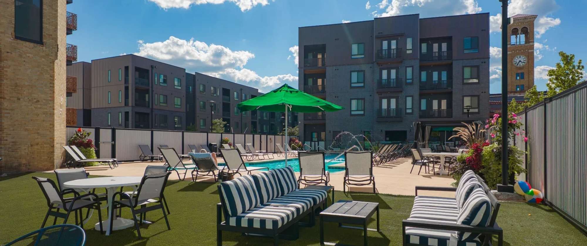 Outdoor pool lounge area at Factory 52 featuring striped seating, green umbrella, patio tables, and apartment buildings with modern balconies in the background.