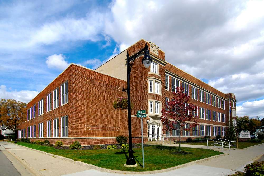 Red brick building with large windows and landscaped entrance on a sunny day at Sycamore House Senior Living in Durand, Michigan