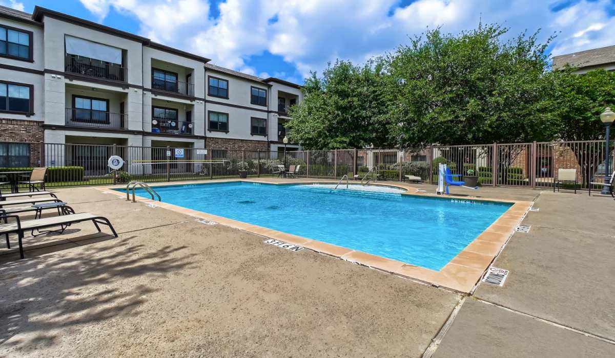 Sparkling pool surrounded by lounge chairs and gated entrance with lush greenery at Lindbergh Parc Senior Apartments in Fort Worth, Texas