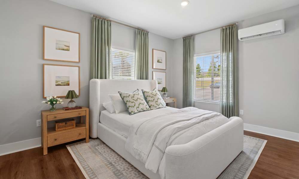 Bright bedroom with large windows, soft bedding, and wood-style flooring, featuring decorative artwork and green curtains at Everett at Port Wentworth in Port Wentworth, Georgia