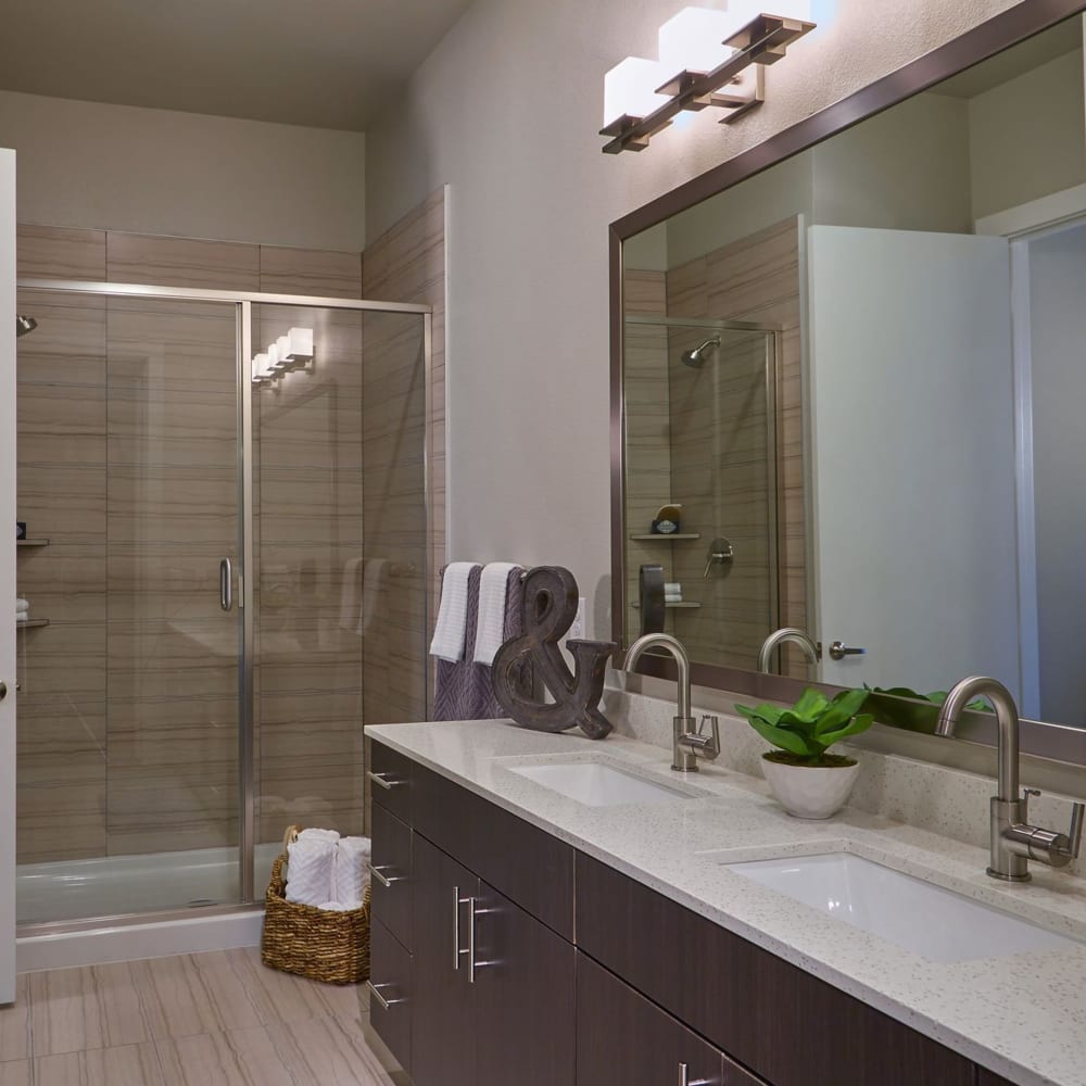 Walk-in shower with white vanity top, large mirror and dark cabinetry at Harvest Lofts in Dallas, Texas