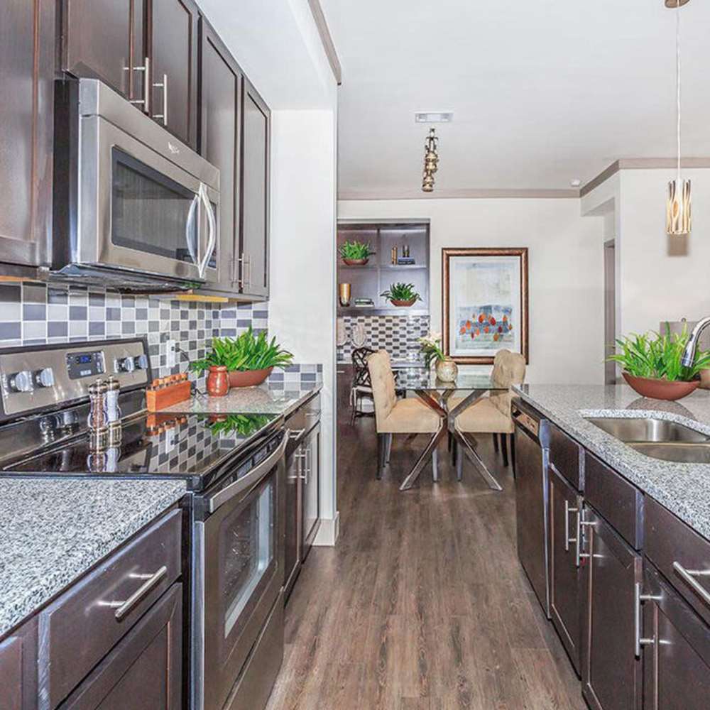 Modern kitchen with granite countertops, stainless steel appliances, and wood-style flooring. Dining area features glass table and stylish chairs at Plantation Park Apartments in Lake Jackson, Texas