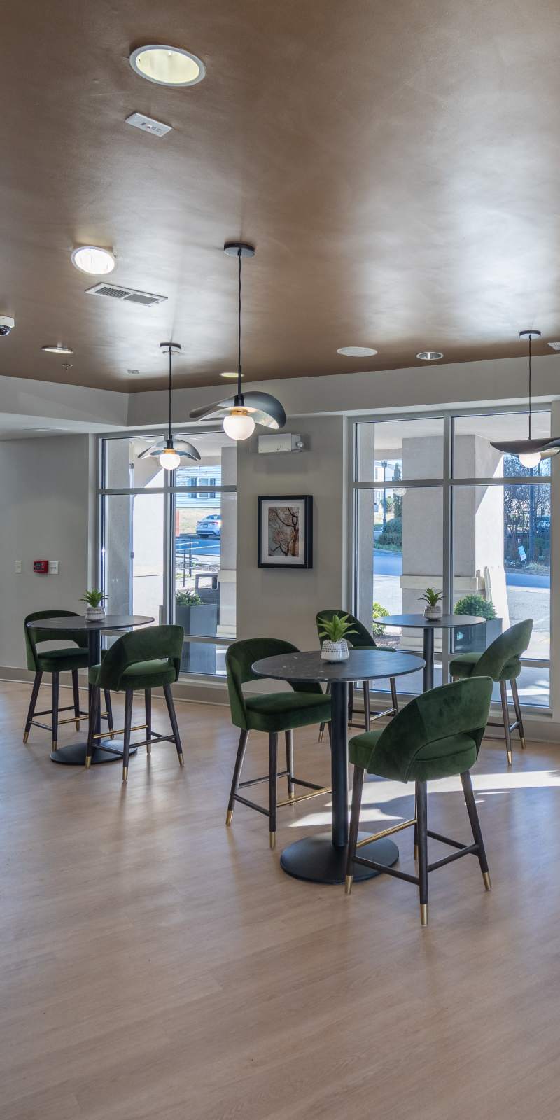 Bright community café-style lounge with round tables, green velvet chairs, modern pendant lighting, and large windows providing views of the outdoor area at The James in Richmond, Virginia