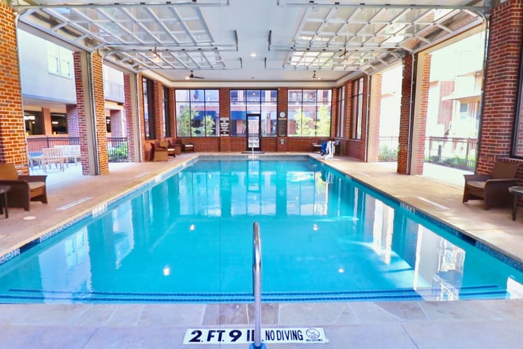 Indoor pool at Watermere Frisco with clear blue water, brick walls, lounge seating, and roll-up garage-style windows at Watermere at Frisco in Frisco, Texas