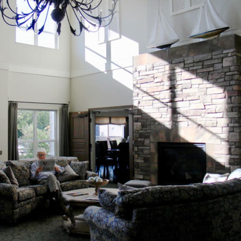 Resident sitting on a couch reading a newspaper in a large fireplace lounge at Deephaven Woods in Deephaven, Minnesota