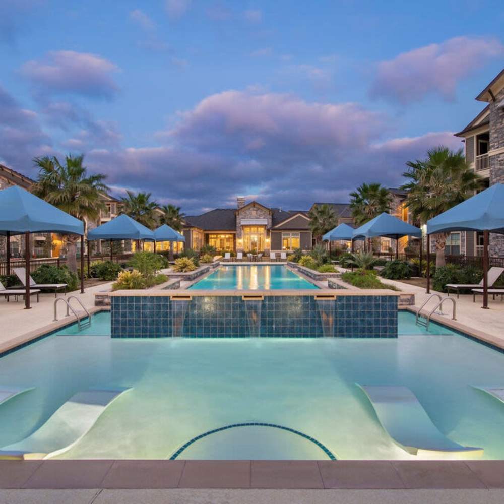 Resort-style sparkling pool surrounded by lounge chairs and shaded cabanas with palm trees at Plantation Park Apartments in Lake Jackson, Texas