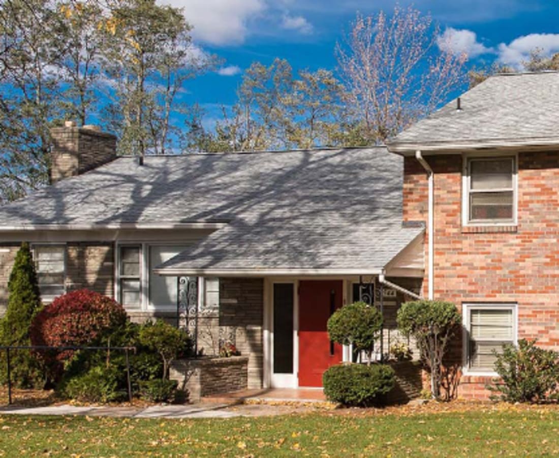 A suburban brick house with a gray roof, red front door, and surrounded by autumn trees under a bright blue sky at Dohr Apartments in Rochester, New York