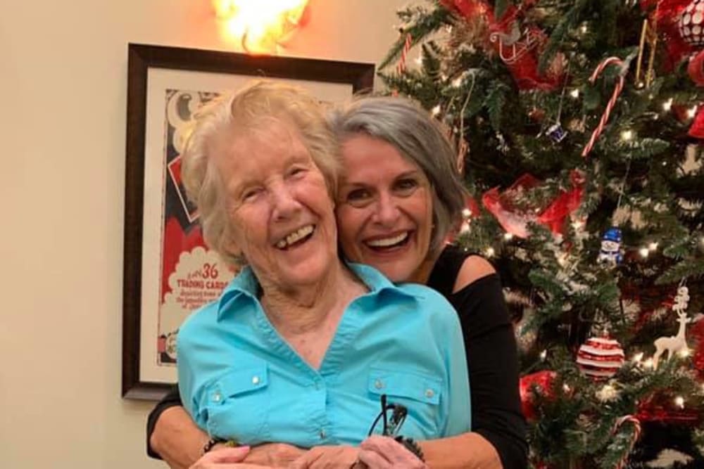 Two resident friends hugging by a Christmas tree at Inspired Living Bonita Springs in Bonita Springs, Florida