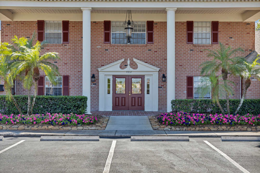 Brick apartment building entrance with white columns, red shutters, landscaped palm trees, and a walkway leading to double doors at Carlton Arms Of Magnolia Valley in New Port Richey, Florida