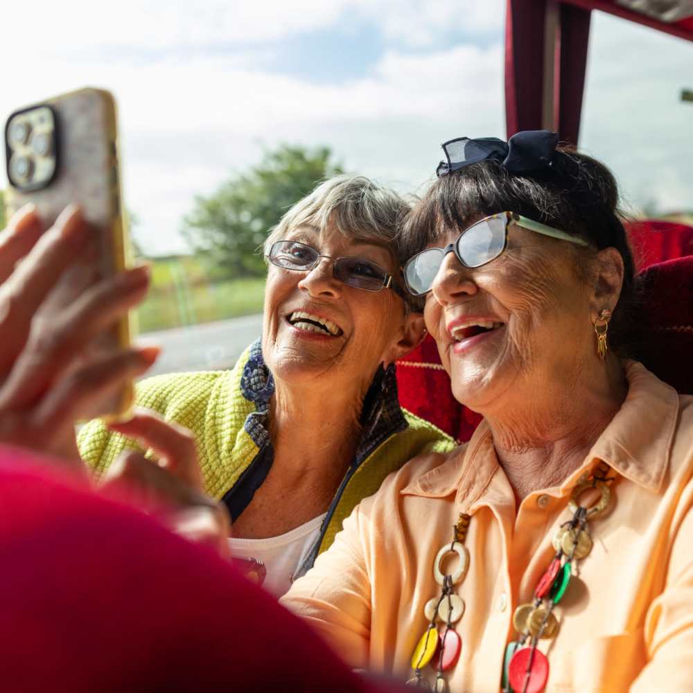 Two smiling older women taking a selfie on a bus, enjoying their time together at Lindbergh Parc Senior Apartments in Fort Worth, Texas