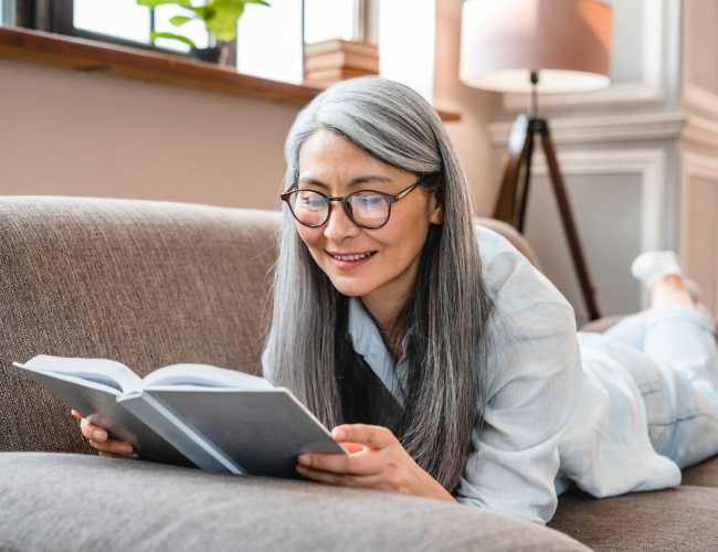 Senior woman reading book by relaxing on sofa at Camino Al Oro in Los Angeles, California