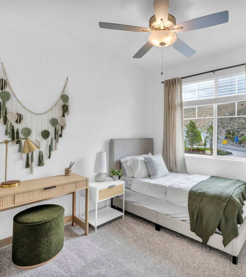 Cozy bedroom with single bed, green throw, nightstand, desk, wall art, beige curtains, and ceiling fan at Prosper 83 in Sparks, Nevada