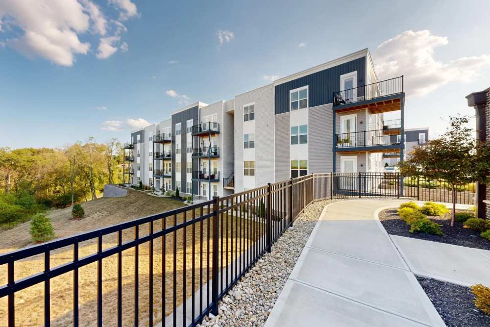 A modern four-story apartment building at Elevation 800 with private balconies, white and dark gray exterior siding, and a landscaped walkway bordered by a black metal fence overlooking a wooded hillside.
