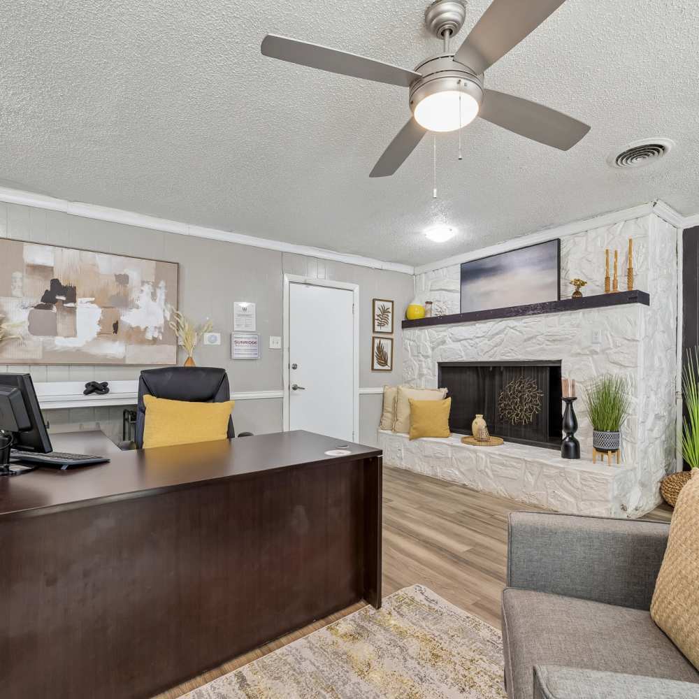 Bright front office and lounge with dark wood desk, gray chair, stone fireplace, ceiling fan, plants, and warm neutral décor at Royal Arms in Richardson, Texas