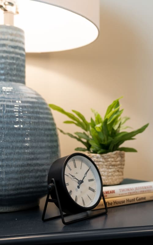 Bedside table with lamp, potted plant, and clock at The Pillars of Hermantown in Hermantown, Minnesota