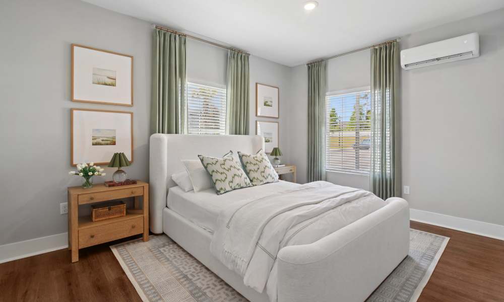 Bright, modern bedroom featuring a white upholstered bed, soft green accent pillows, floor-to-ceiling green curtains, wood nightstand, and large windows with natural light.