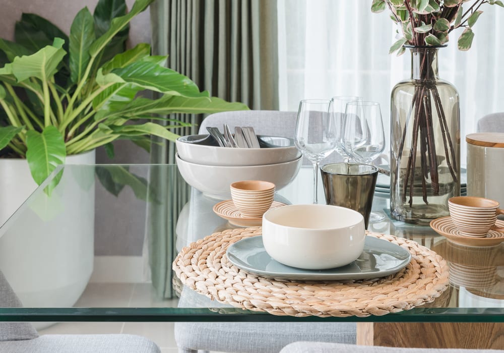 Glass kitchen table with a place setting of two tea cups, a ceramic bowl on top of a dinner plate, two large stacked bowls with silverware placed inside, a vase and flowers on top and a large plant in the background at Everwood at The Avenue in Murfreesboro, Tennessee