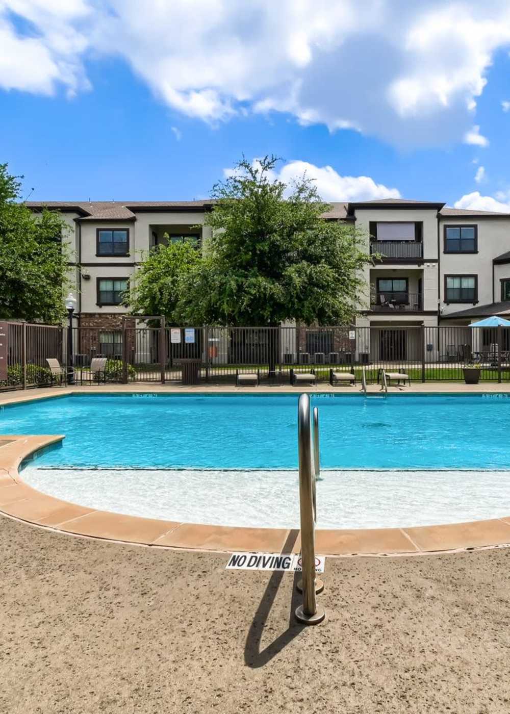 Sparkling pool surrounded by lounge chairs and trees with a gated entrance in the background at Lindbergh Parc Senior Apartments in Fort Worth, Texas