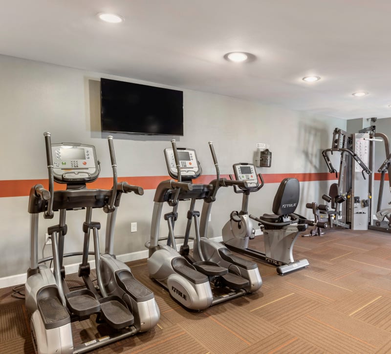 Cardio machines in front of a wall-mounted TV screen in the fitness center at Foundations at Austin Colony in Sugar Land, Texas