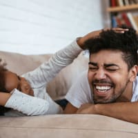 Resident family consisting of dad and child wrestling, laughing and smiling together on a couch in living room at Decker Apartment Homes in Ft Worth, Texas