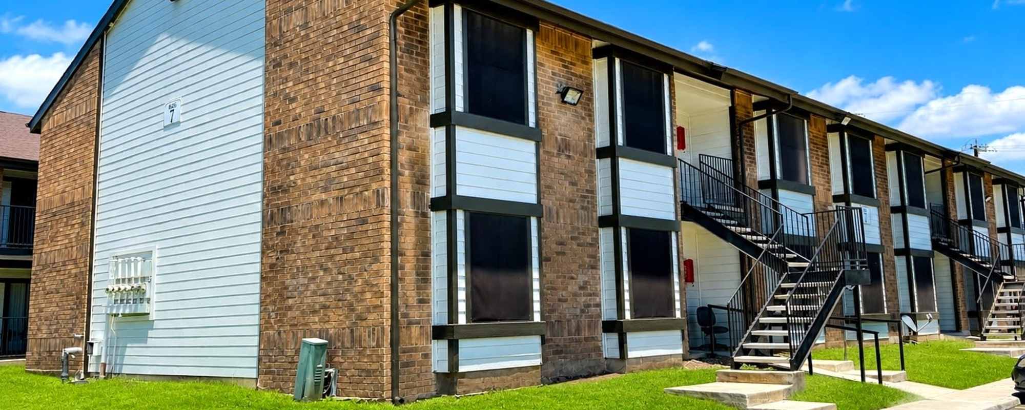 Image of a freshly painted apartment building with brick siding and staircases in front leading to different entry doors in a bright blue sky at Towne East Village Apartment Homes in Converse, Texas