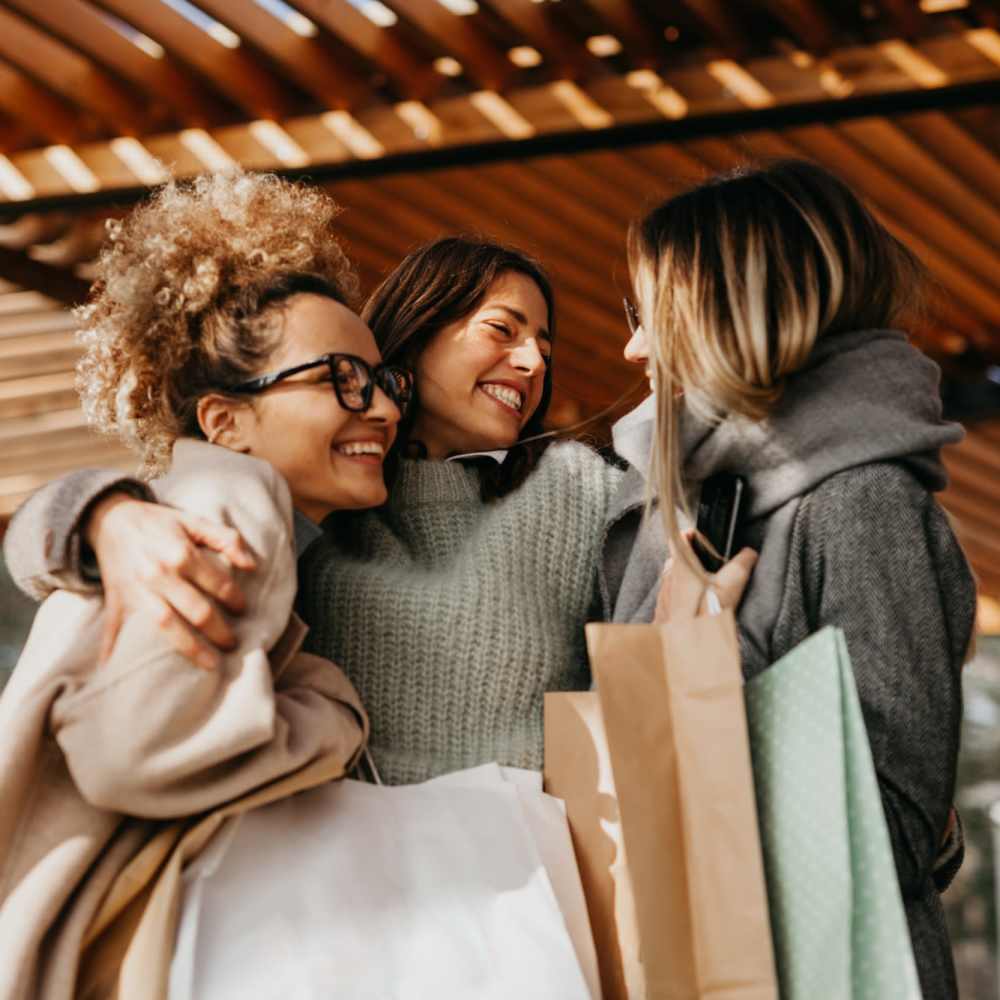 Three women smiling and embracing, holding shopping bags under a wooden pergola at The Arcadia in Madison, Tennessee