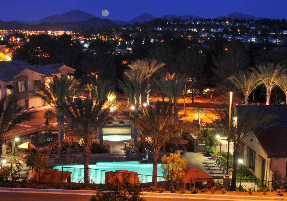 Breathtaking night view showcasing a sparkling water pool surrounded by lush landscaping and softly glowing palm trees, framed by the scenic hills and twinkling city lights at Cresta Bella in San Diego, California