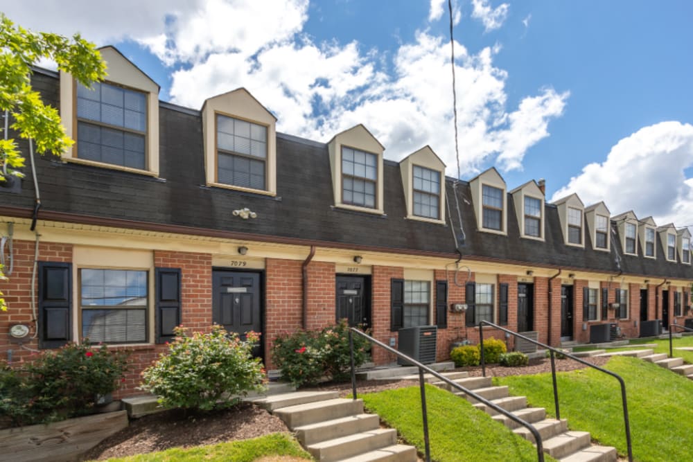 Row of townhomes with brick facades, lush landscaping, and clear blue sky overhead at Dutch Village Apartments in Baltimore, Maryland