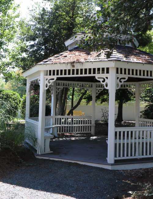A white wooden gazebo with brown shingled roof, surrounded by trees and greenery at The Crossings at Ironbridge in Chester, Virginia