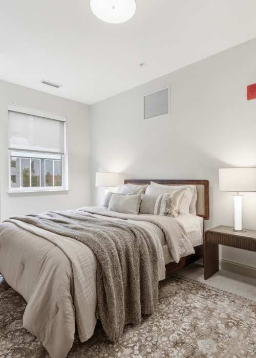 Cozy bedroom with layered blankets, soft carpet, and large window view at The Lawrence in Buffalo, New York