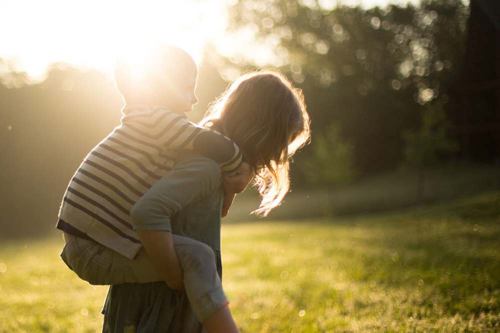 Resident carrying young son on her back as they walk to the playground across the beautifully green grass at sunrise with a silhouette of trees behind them at Eagle Point Village in Fayetteville, North Carolina