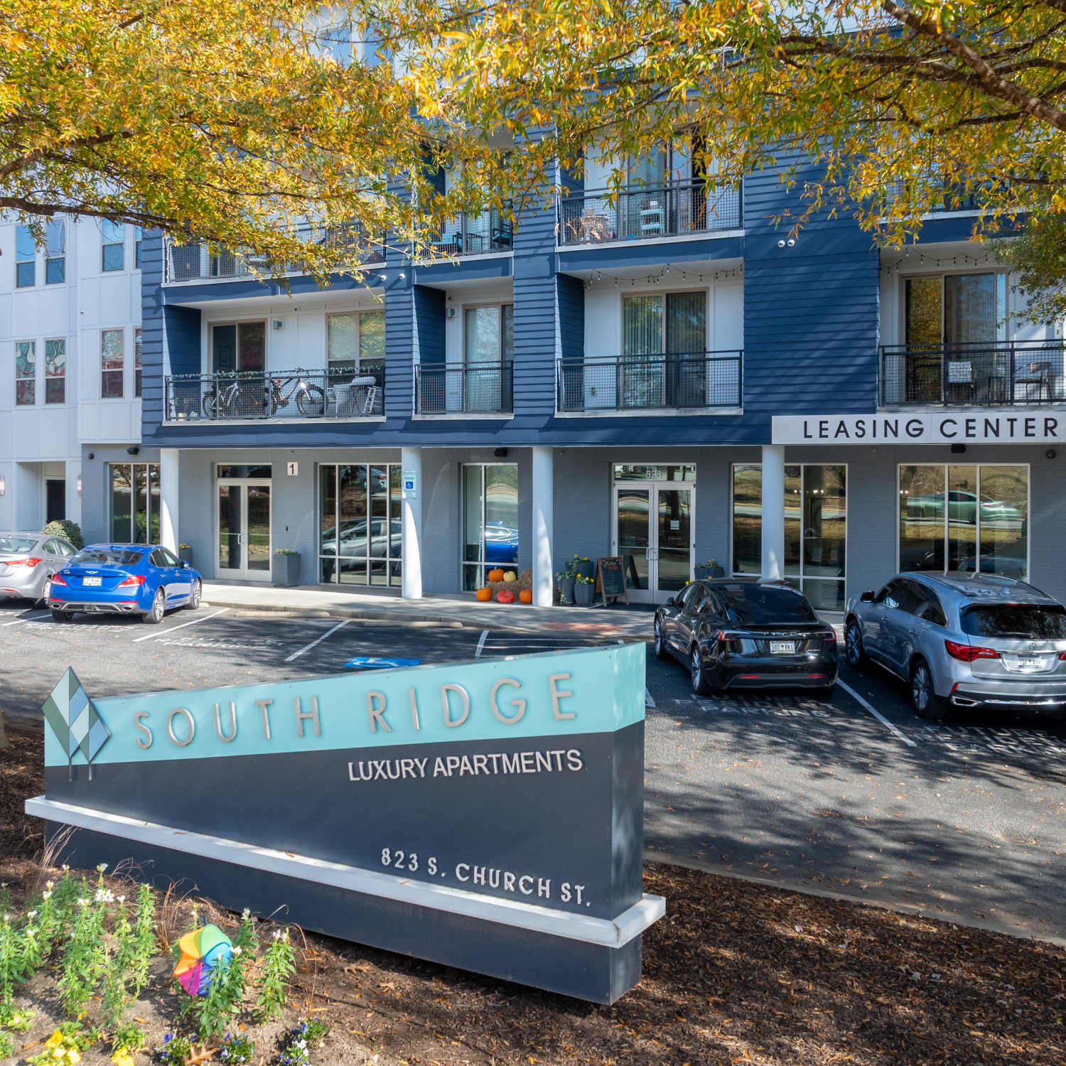 Modern apartment building with blue-and-white facade, leasing center sign, and autumn trees in front in South Ridge in Greenville, South Carolina