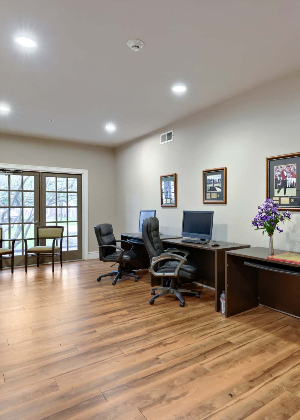 Business center with computer stations, ergonomic chairs, wood-style flooring, and large glass doors leading outside at Lindbergh Parc Senior Apartments in Fort Worth, Texas