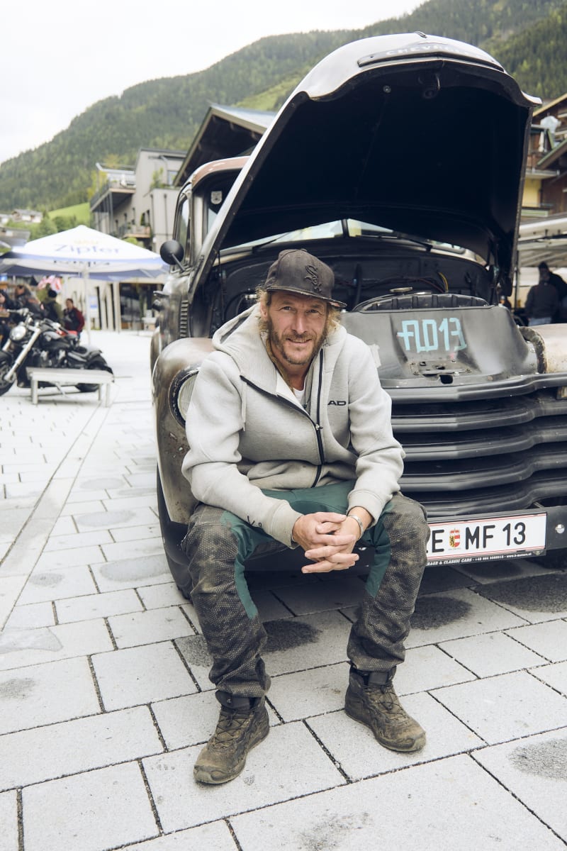 Ein Mann, Flo Wolf sitzt vor einem restaurierten Pickup im Biker Village der Biker Mania reloaded. Die Umgebung zeigt alpenländische Architektur.