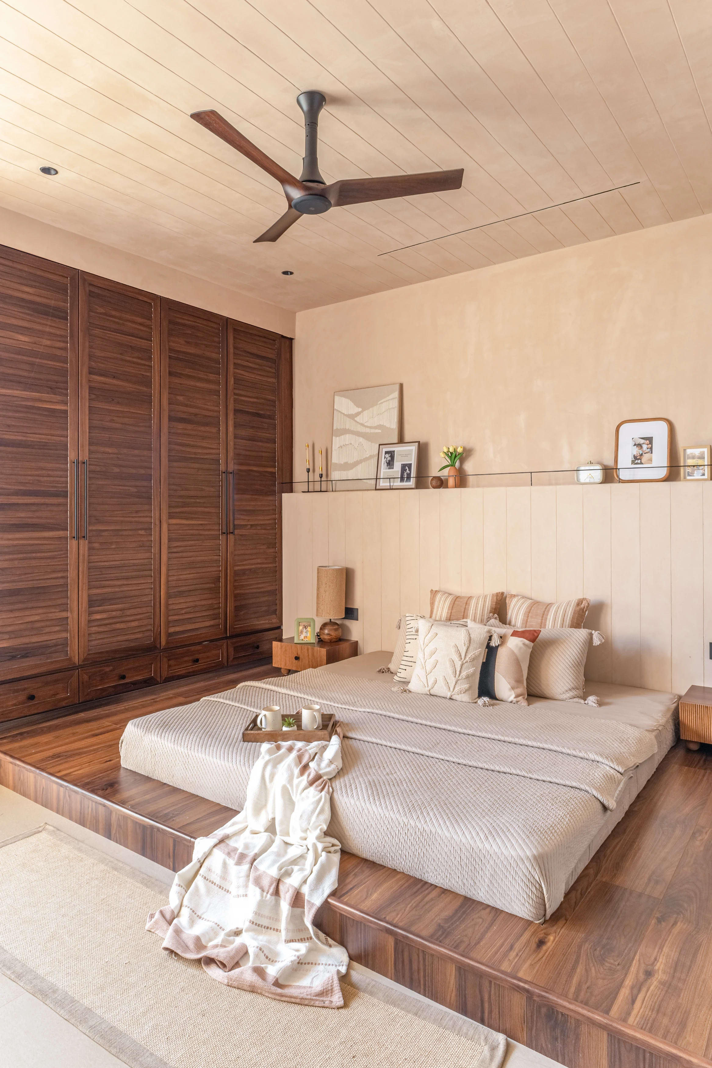 Bedroom in Sukoon: a platform bed on a raised wood plinth against a full-height walnut wardrobe wall, under a timber-clad ceiling
