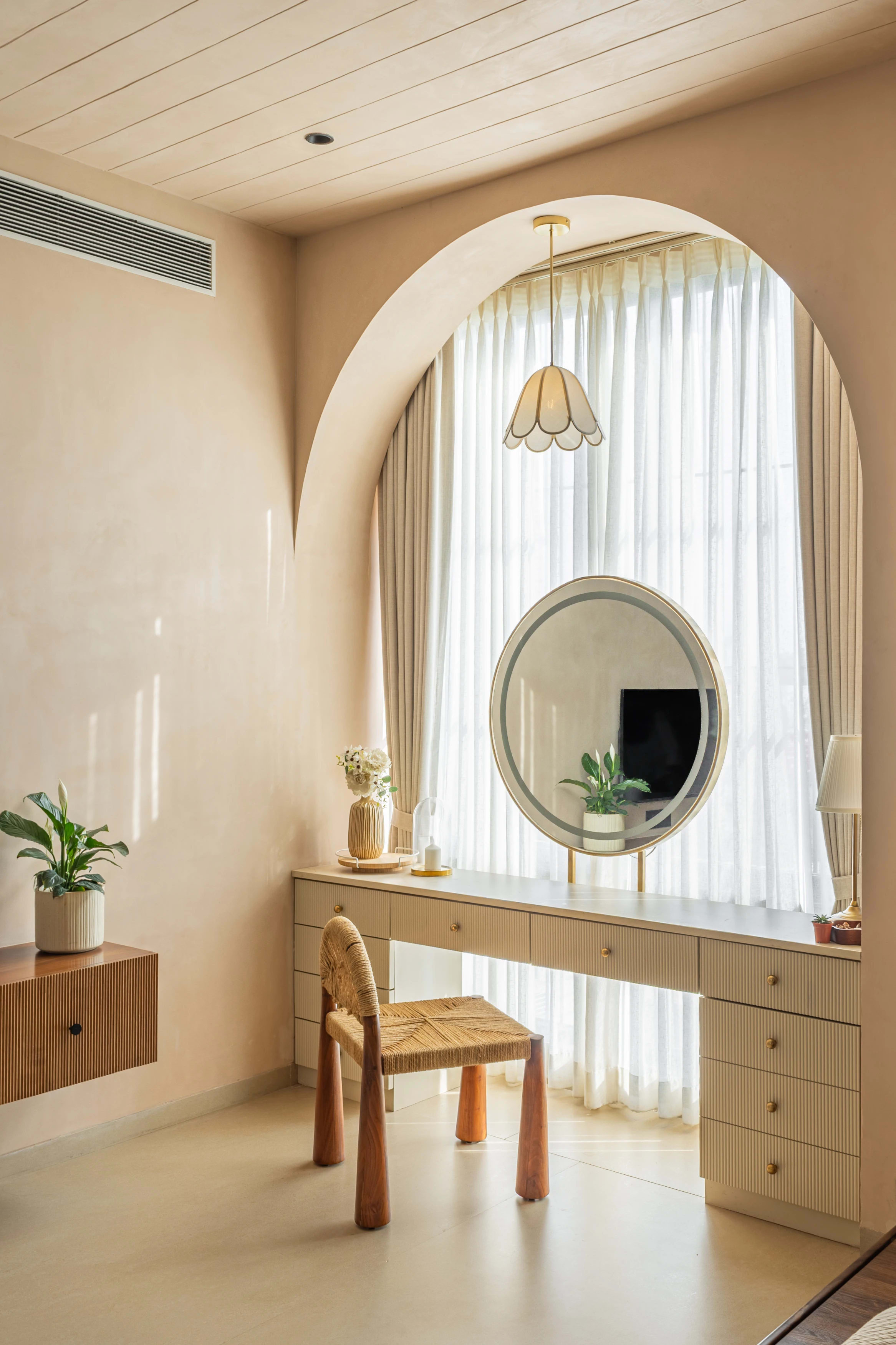 Dressing area in Sukoon: a fluted cream dressing table set into an arched lime-plaster alcove, with a round backlit mirror, a petal pendant and a cane-seated chair